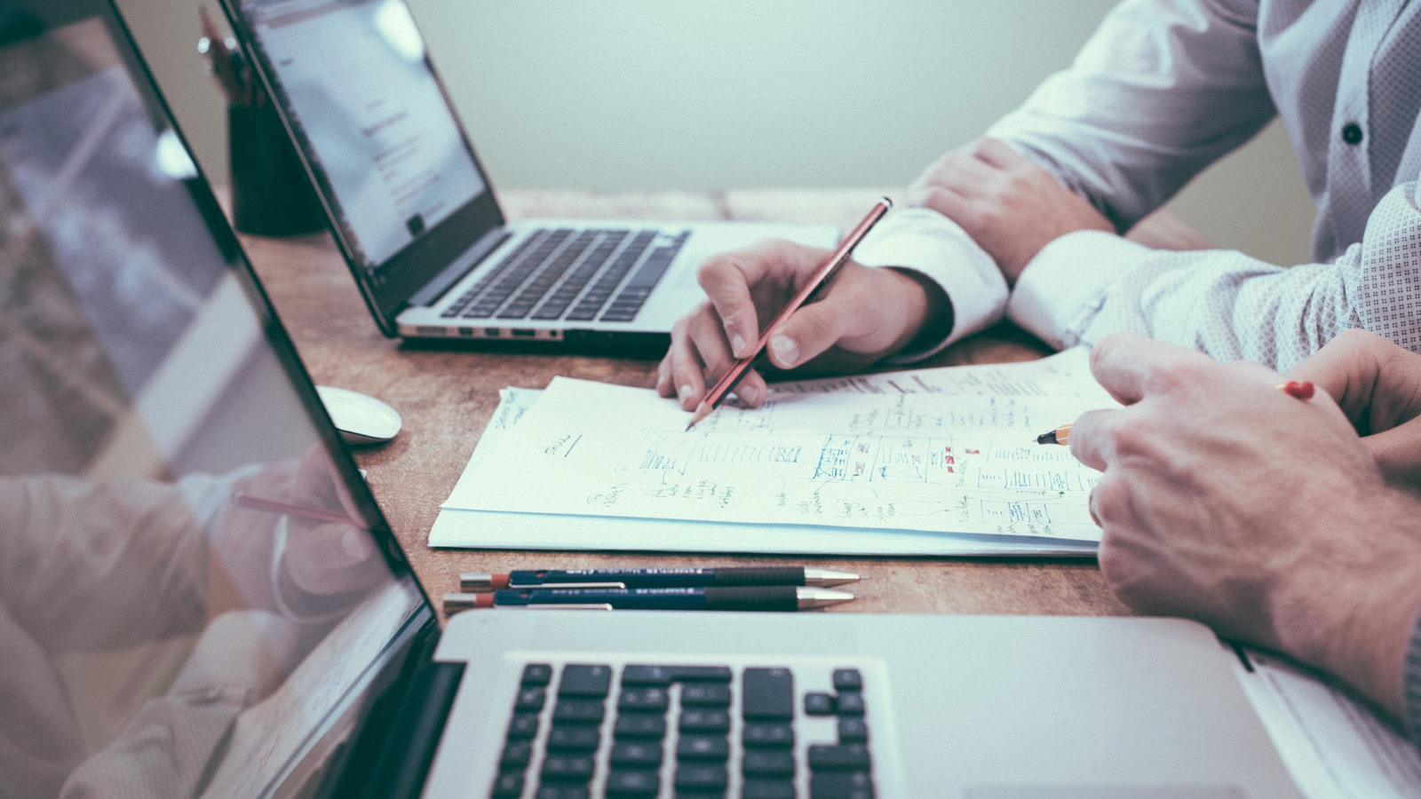 Two people compare handwritten ideas on paper beside open laptops.