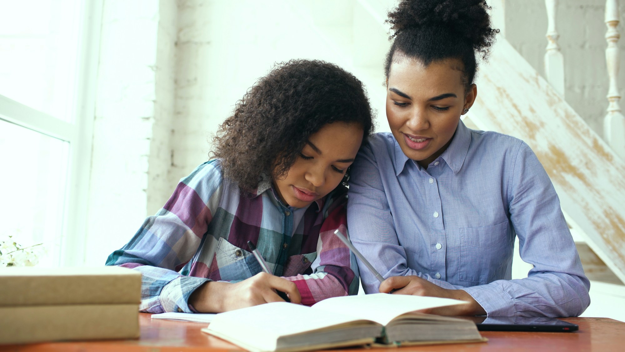 A mother and teenage daughter review a notebook together with a smartphone resting on the table.